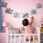 A baby in a pink outfit stands in a white crib, reaching up at a string of personalized unicorn and rainbow clip-art cards hanging on a pink wall.